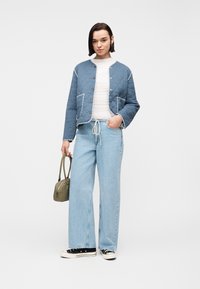 Γυναίκα με γαλάζιο καπιτονέ μπουφάν, λευκό τοπ, ανοιχτό μπλε τζιν με φαρδιά μπατζάκια, κρατώντας μια ελιά τσάντα και φοράει μαύρα αθλητικά παπούτσια.