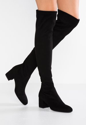 Bottes noires au-dessus du genou avec talons blocs bas portées par une personne se tenant sur une surface blanche.