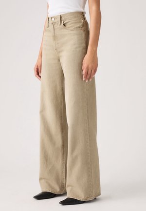 Vrouw die beige broeken met hoge taille en wijde pijpen draagt, gecombineerd met een wit topje en zwarte puntige schoenen, staand tegen een effen achtergrond.