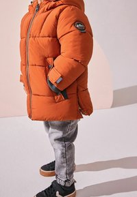 Doudoune orange avec capuche, fermeture éclair et poches latérales ; présente des accents gris et un logo circulaire ; portée avec un pantalon de survêtement gris et des baskets noires.