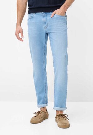 Mand iført lyseblå opkrogede jeans og brune snørebåndssko, står med den ene hånd i lommen på hvid baggrund.