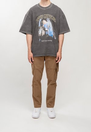 Maglietta grigia oversize con grafica di leone e angelo, abbinata a pantaloni cargo color cachi. Completano il look le sneakers bianche.