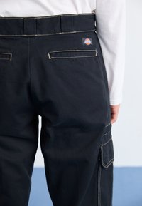Black cargo pants with yellow stitching, two back pockets, and a side cargo pocket, made of durable fabric. Features brand logo patch.
