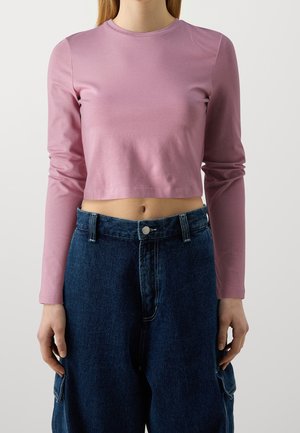 Vrouw met een roze cropped top met lange mouwen en een hoge taille donkerblauwe spijkerbroek met cargozakken tegen een effen lichte achtergrond.