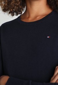 Person som bär en mörk marinblå tröja med rund hals och en liten röd, vit och blå logotyp på bröstet, med armarna i kors.