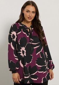 Blusa a maniche lunghe con scollatura arricciata e dettagli con fiocco. Base viola scuro con grandi motivi floreali neri e rosa; tessuto testurizzato.
