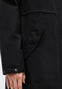 Fekete kabát ujja és zsebe közelről, patentos zsebfedéllel, amelyet egy, a csuklójánál látható kézzel visel egy személy.