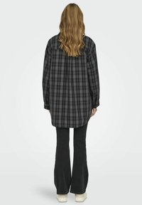 Camisa de cuadros en negro y gris, de manga larga, con corte relajado y dobladillo curvado, combinada con pantalones de campana negros y zapatos de color claro.