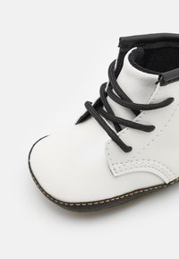 Bota de cuero blanca con cordones negros, punta redonda y costuras amarillas a lo largo de la suela. Textura suave con una parte superior ligeramente acolchada.