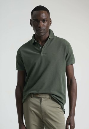 Polo de color verde oscuro con cuello, mangas cortas y tapeta de dos botones. Tejido texturizado con sutil logo de la marca. Combinado con pantalones beige.
