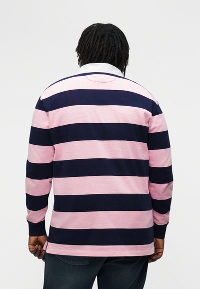 Mann iført rugbygenser med lange ermer og brede, horisontale marineblå og rosa striper, hvit krage, sett bakfra mot ensfarget bakgrunn.