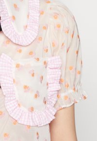 Tissu blanc transparent avec broderies florales pêche et orange, détails en vichy rose, col à volants et manches bouffantes. Comprend une poche.