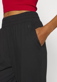 Černé kalhoty s elastickým pasem, bočními kapsami a měkkou, texturovanou látkou. Na boku je viditelný detail loga.