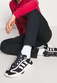 Svartvita sportskor med rund design, textilstruktur, tre randiga accenter och en kraftig sula. Bärs med vita strumpor.