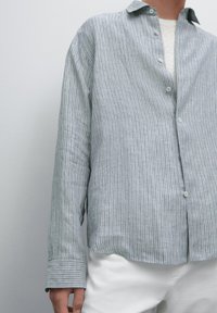 Person wearing a light gray vertically striped button-up shirt over a white top and white pants against a plain background.