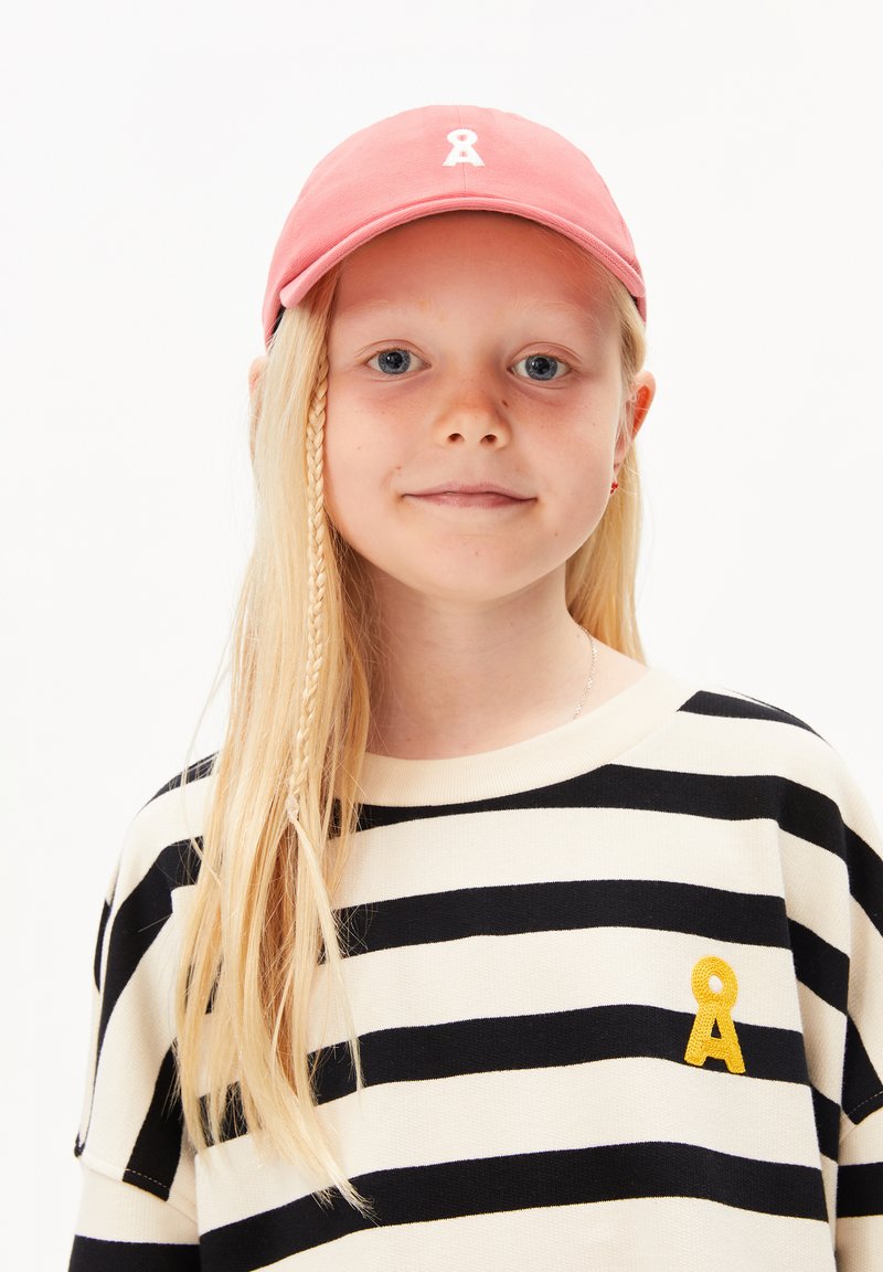 Casquette rose avec logo en ruban blanc, associée à un sweatshirt rayé crème et noir avec un détail brodé jaune. Cheveux longs et clairs.