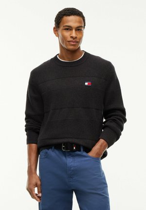 Junger Mann, der einen schwarzen Strickpullover mit kleinem Logo und blauen Hosen trägt, steht mit einer Hand in der Tasche vor weißem Hintergrund.