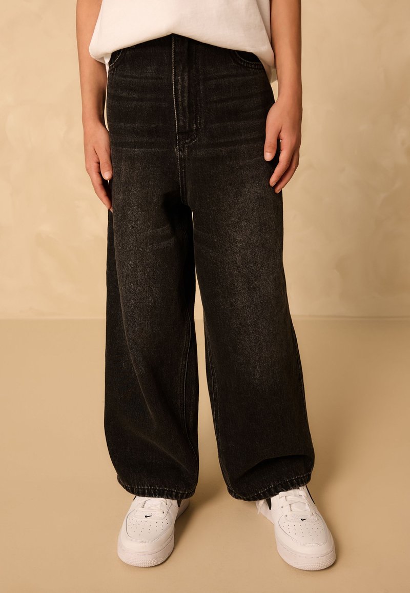 Personne portant un jean noir ample et des baskets blanches, debout devant un fond beige uni.