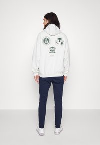 Sudadera blanca con capucha y múltiples logotipos en verde oscuro en la parte posterior, combinada con pantalones de chándal azul marino y zapatillas bajas blancas.
