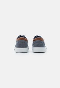Baskets en tissu bleu avec un accent en cuir marron au talon et une semelle en caoutchouc blanc, présentant un design low-top et un tissu texturé.