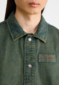 Jachetă din denim verde cu guler ascuțit, având o patch bleu cu text alb. Include închidere cu capse metalice și țesătură texturată.