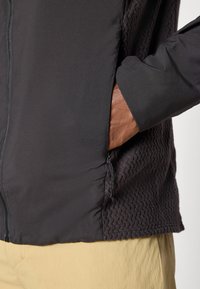 Käsi musta vetska-jaki küljetaskus, millel on tekstureeritud kangapanellid, kantud üle beežide pükste ja ühevärvilise heleda tausta ees.