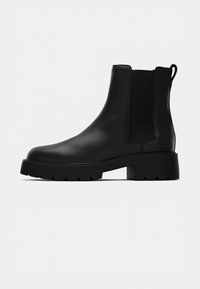 Bota de tornozelo em couro preto com bico arredondado, painéis elásticos e uma sola de borracha robusta. Possui uma aba para facilitar o calce.