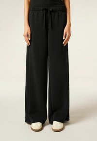 Zwarte wide-leg broek van zachte stof met een elastische tailleband en een trekkoorddetail, gecombineerd met witte instap schoenen.