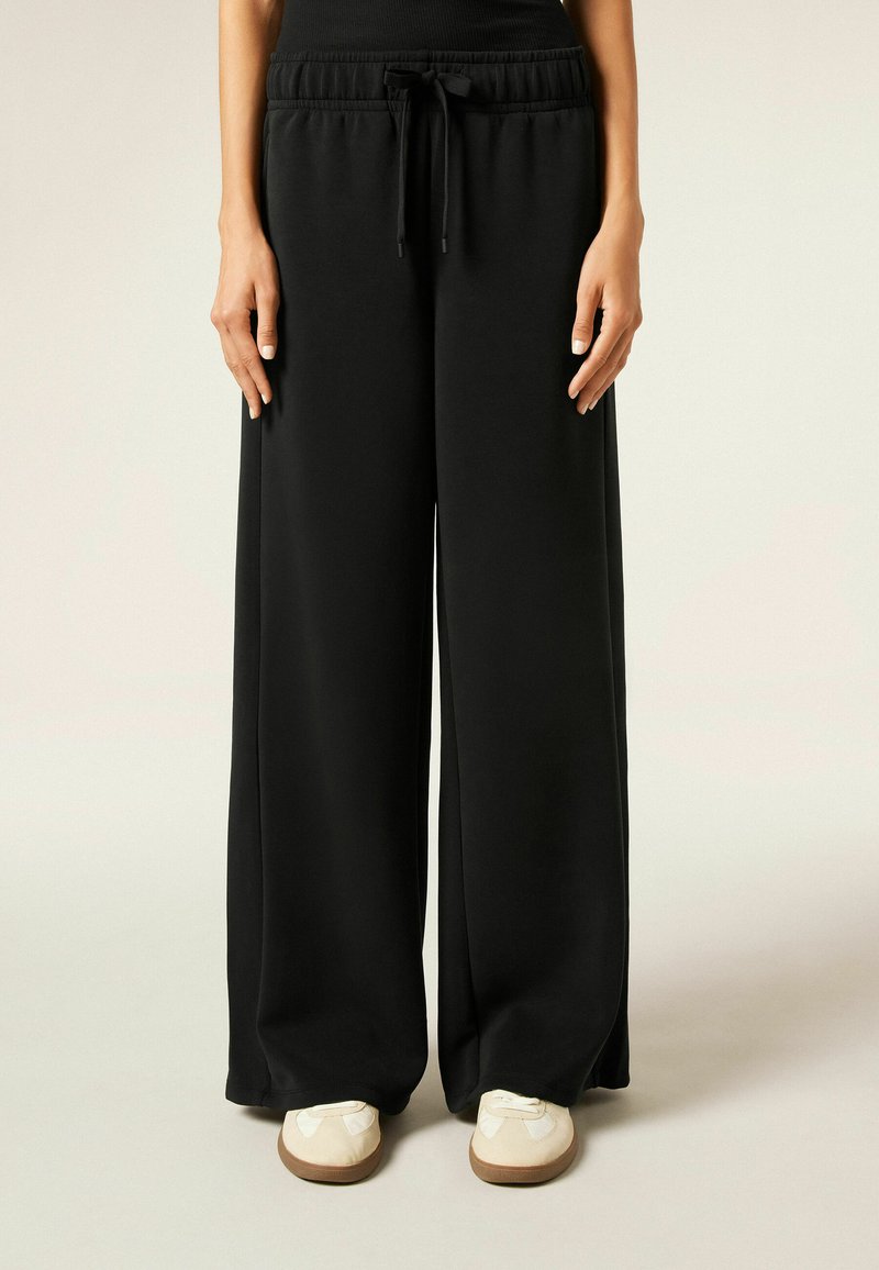 Zwarte wide-leg broek van zachte stof met een elastische tailleband en een trekkoorddetail, gecombineerd met witte instap schoenen.