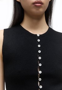 Młoda osoba nosząca czarny bezrękawnik z pionowym rzędem białych guzików z przodu, niektóre rozpięte.