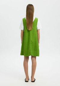 Vestido de lino verde con un diseño sin mangas, que cae por encima de la rodilla. Camiseta blanca como capa inferior visible en el escote. Líneas simples y limpias.