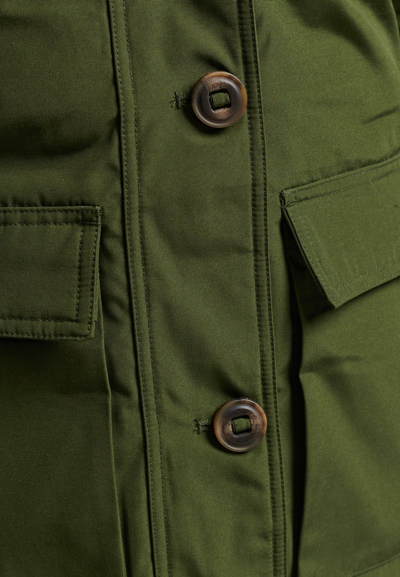 Olivgrüne Jacke mit Knopfverschluss, ausgestattet mit aufgesetzten Taschen und sichtbaren Nahtdetails. Glatte, matte Stoffoberfläche.