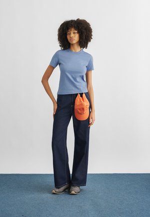 Joven con cabello rizado que lleva una camiseta azul, jeans amplios oscuros, zapatos grises, sosteniendo una gorra naranja frente a sus pantalones.