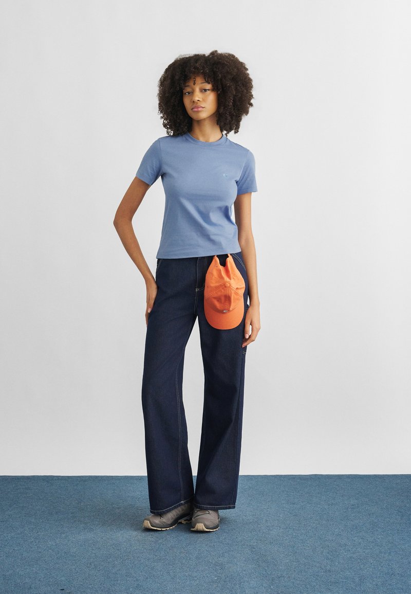 Νέα γυναίκα με σγουρά μαλλιά, που φοράει μπλε μπλούζα, σκούρα τζιν με φαρδιά πόδια, γκρι παπούτσια και κρατάει ένα πορτοκαλί καπέλο μπροστά από το παντελόνι της.