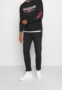 Sudadera negra con el texto "DISILLUSION" y detalles en rosa en la manga, combinada con jeans negros y zapatillas blancas.