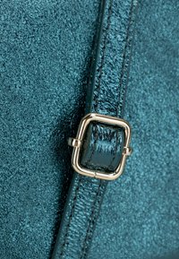 Borsa in pelle testurizzata color teal, in primo piano, con una fibbia in metallo dorato e cuciture, che mostra una finitura liscia e un colore ricco.