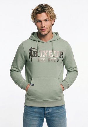 Joven con cabello rubio que lleva una sudadera con capucha verde clara con el texto y gráfico "Boxeur des Rues", manos en el bolsillo delantero, vaqueros azules.