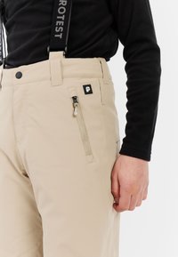 Beige ski-broeken met ritszak, verstelbare bretels en een zwart logo-patch aan de zijkant. Materiaal lijkt glad en duurzaam.