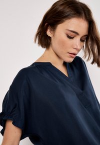 Navy blue, lightweight blouse with a V-neckline and short puff sleeves. Smooth texture with subtle sheen and a small front placket.