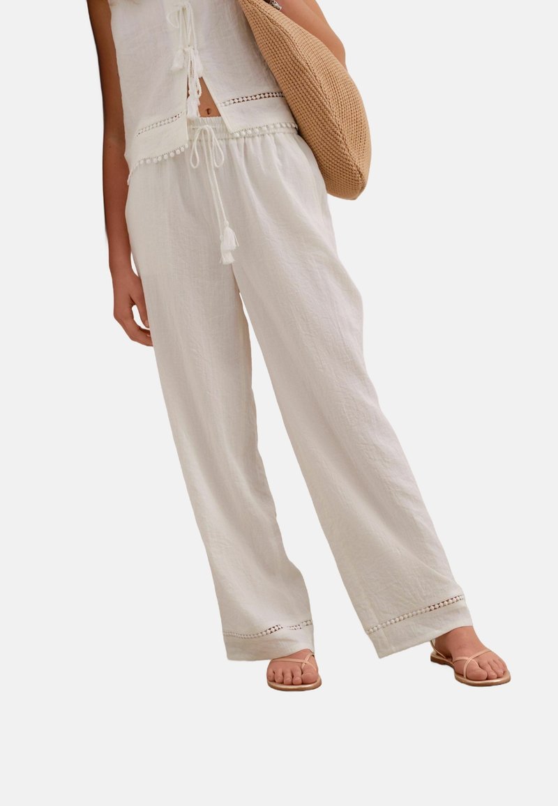 Femme portant un pantalon blanc ample et un haut sans manches, portant un sac en tissu tressé beige et des sandales beiges.