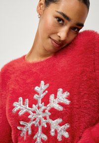 Rotem, flauschiger Pullover aus weichem Material, mit einem silbernen Pailletten-Schneeflockenmotiv in der Mitte. Rundhalsausschnitt und lockere Passform.