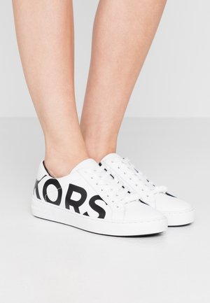 Person iført hvide lavtop-sneakers med sort fed skrift på siderne, stående på en lysegrå gulv.