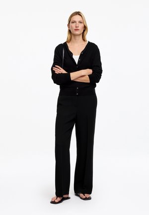 Femme aux cheveux blonds debout, bras croisés, portant un cardigan noir, un pantalon noir à jambes larges, des sandales noires et un haut blanc sur fond blanc.