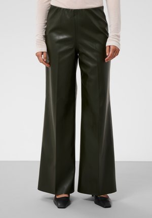 Personne portant un pantalon en cuir vert foncé taille haute à jambes larges, un haut beige à manches longues et des chaussures noires pointues.