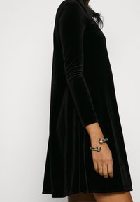 Zwarte fluwelen jurk met lange mouwen en een uitlopende zoom, gecombineerd met een zilveren armband met twee sferische accenten. Gladde textuur overal.