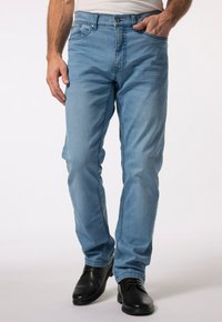 Světle modré džínové kalhoty slim fit s pěti kapsami a zipovým zavíráním. Oblečené s černými lesklými botami, které mají hladkou texturu.