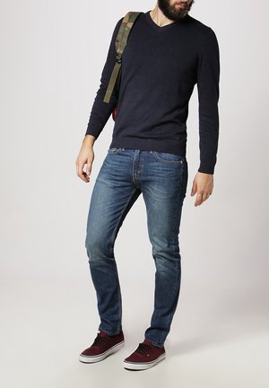 Donkerblauwe v-hals trui, slim-fit blauwe jeans, kastanjebruine sneakers en een rugzak met camouflagepatroon. Casual outfit met contrasterende kleuren.