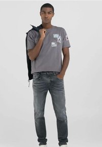 Camiseta de algodón gris con mangas cortas y varios parches, combinada con unos jeans gris oscuro. El modelo sostiene una chaqueta negra sobre un hombro.