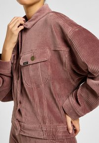 Giacca di velluto a coste rosa con colletto, chiusura con bottoni e due tasche sul petto. Presenta coste verticali e una vestibilità rilassata.