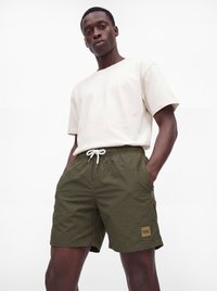 Mann trägt ein schlichtes weißes T-Shirt und olivgrüne Shorts mit Kordelzug, die Hände in den Taschen, vor weißem Hintergrund.